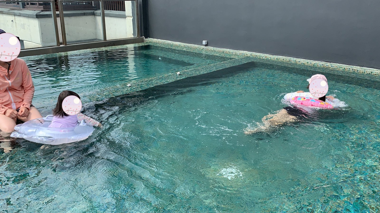Reaching out to my child floating on a swim ring by the pool, another child swimming in the background 
